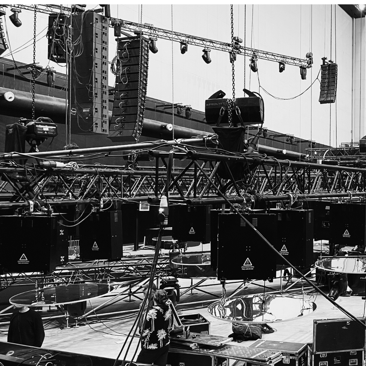 Stage equipment setup featuring suspended lighting and sound systems, with technicians preparing for an event in a large venue. The image showcases a complex arrangement of trusses, speakers, and reflective surfaces on the stage.