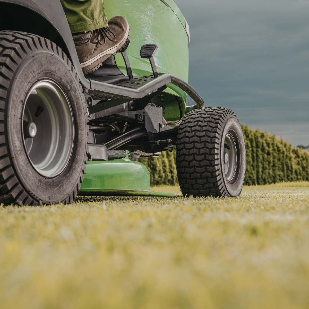 Close-up view of a green lawn mower with a focus on the rear wheel and operator's foot, set on a lush grassy field, highlighting the Auto Lok™ feature for enhanced stability and control during mowing.