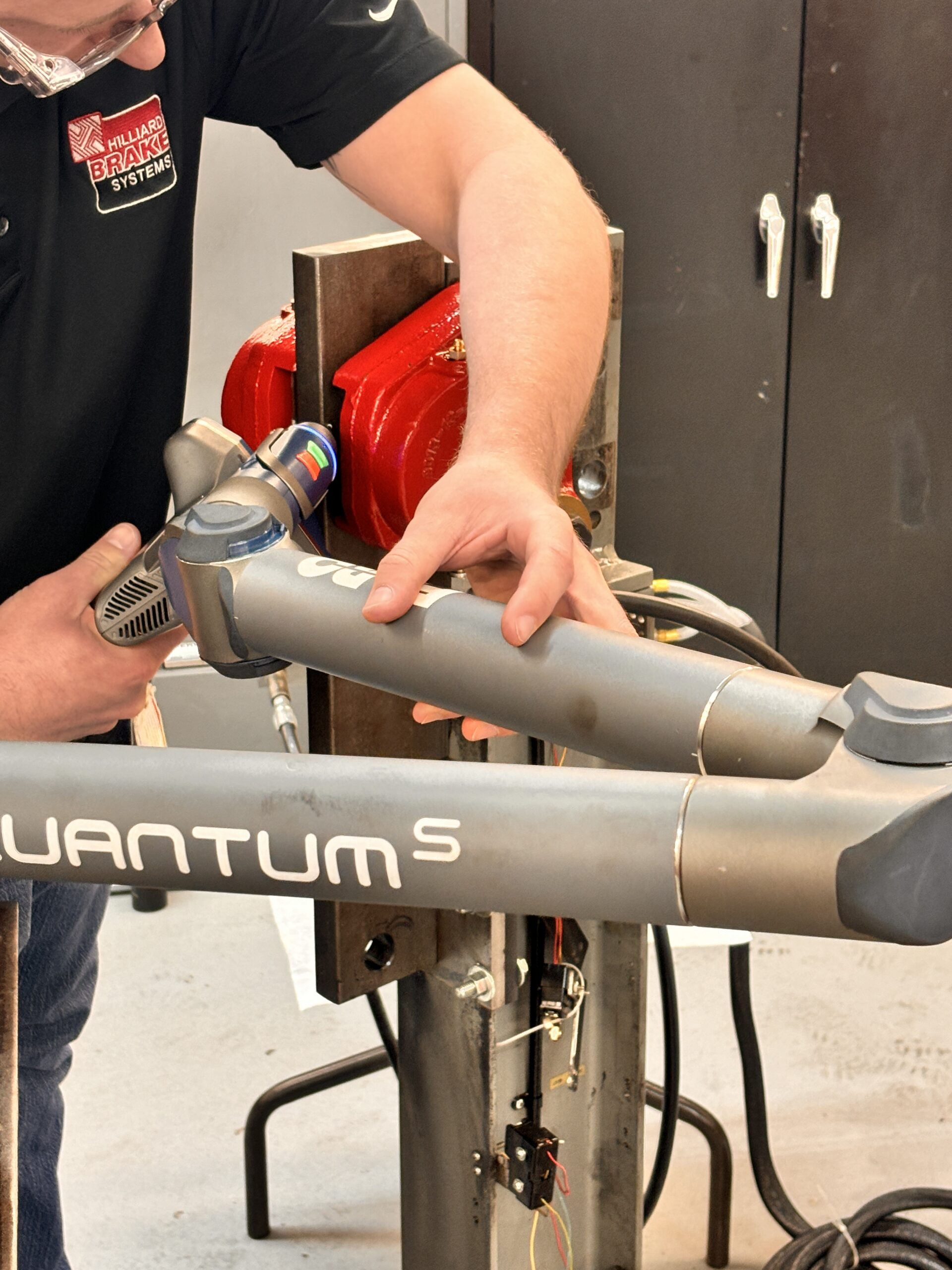 Technician performing maintenance on a bicycle frame in a workshop, using a precision tool on the QUANTUM S model. Red hydraulic equipment is visible in the background, highlighting a professional setting.