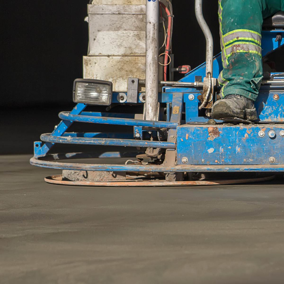 Worker operating a blue concrete finishing machine on a construction site, smoothing freshly laid concrete.
