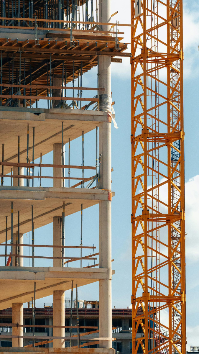 Construction site featuring a partially built concrete structure with scaffolding and an orange crane against a blue sky.