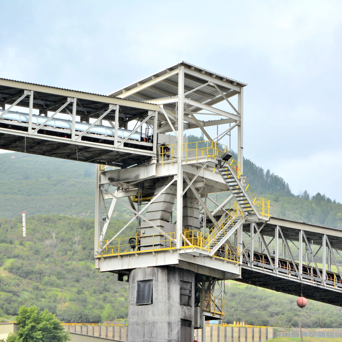 Industrial conveyor system with a steel structure and stair access, transporting materials over a hillside, showcasing efficient bulk handling solutions in a manufacturing environment.