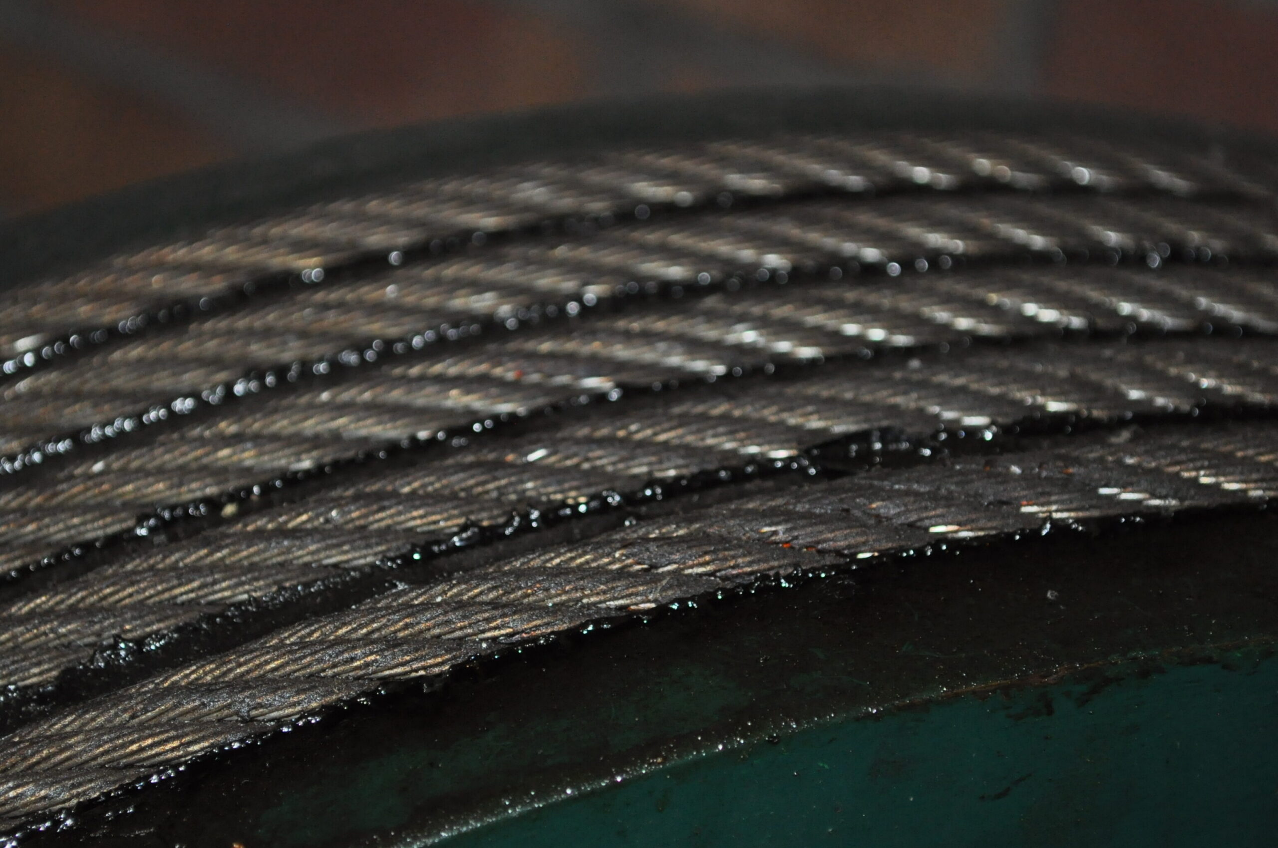 Close-up image of a metal cable wrapped around a large spool, showcasing the texture and sheen of the cable strands against a dark green background. The blurred brick wall in the background adds depth to the industrial setting.