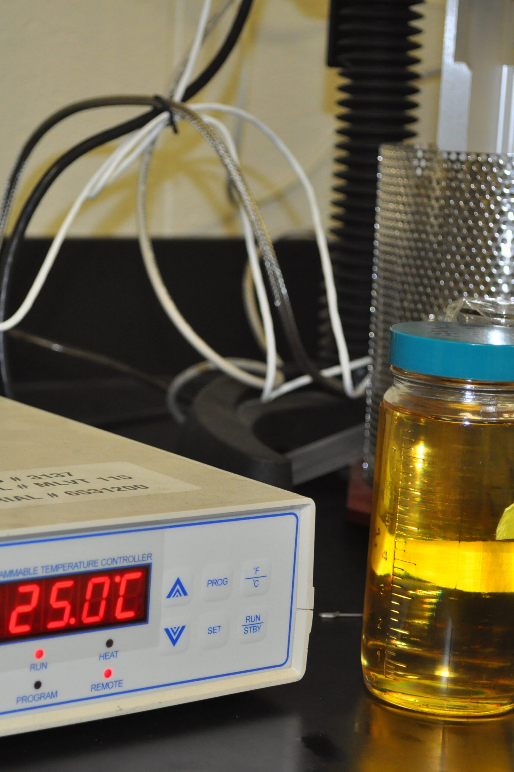 Programmable temperature controller set at 25°C next to jars of fluid samples for analysis, including labeled containers on a laboratory countertop.