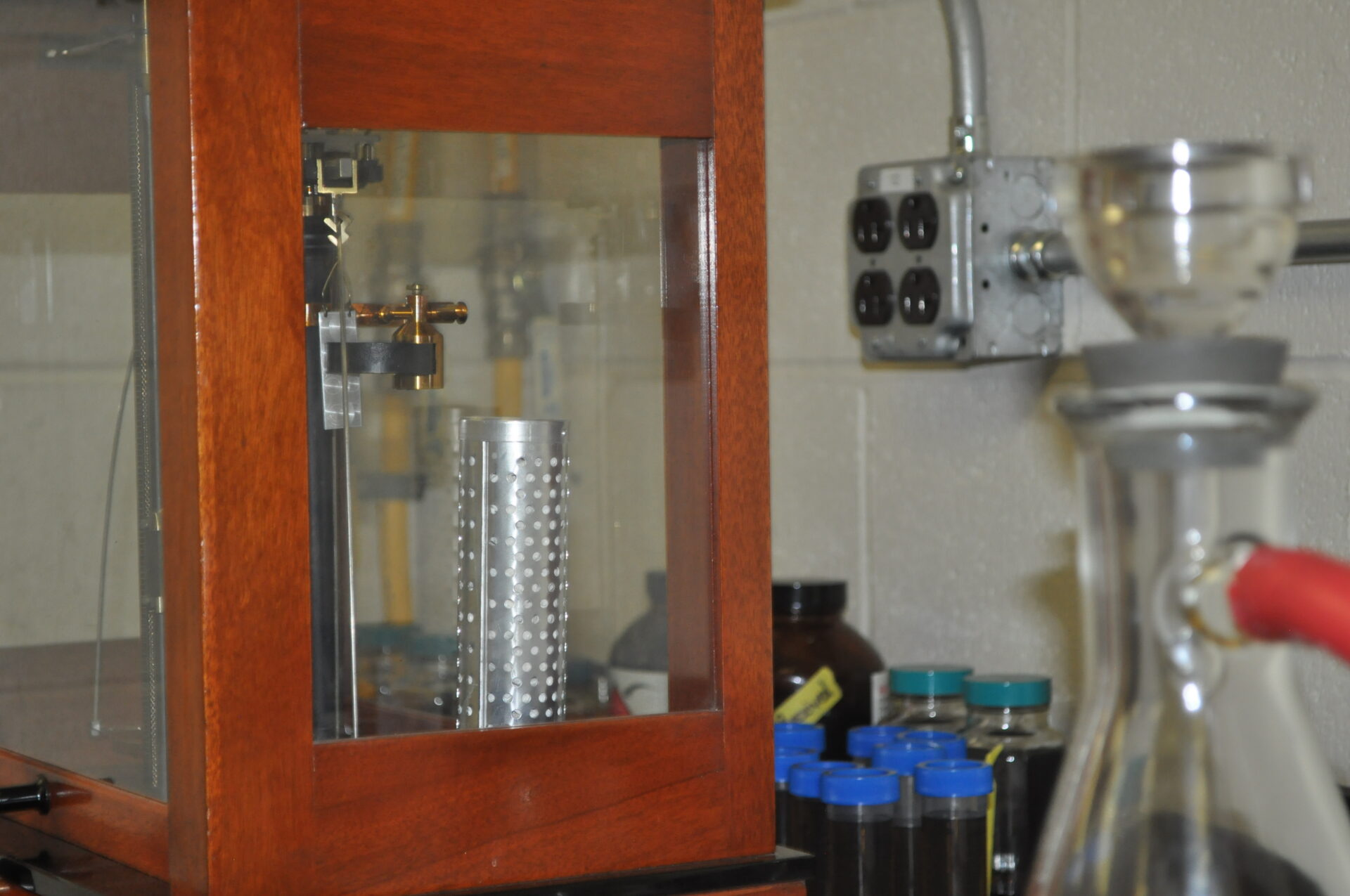 Laboratory equipment showcasing a glass chamber with a metallic cylinder, used for conducting experiments and analysis in lab services. Visible background includes various labeled bottles and electrical outlets, emphasizing a professional scientific environment.