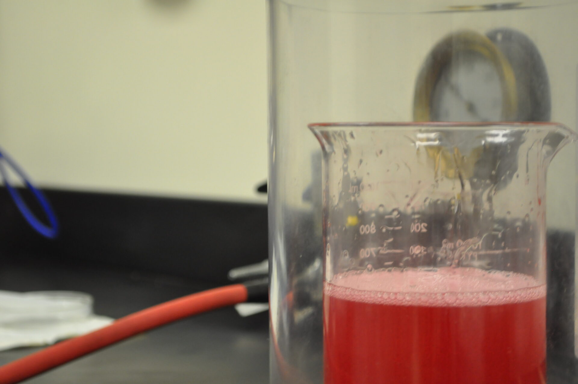 Laboratory beaker containing a red fluid sample, with measurement markings visible, on a lab countertop with a pressure gauge in the background, illustrating fluid sample analysis.