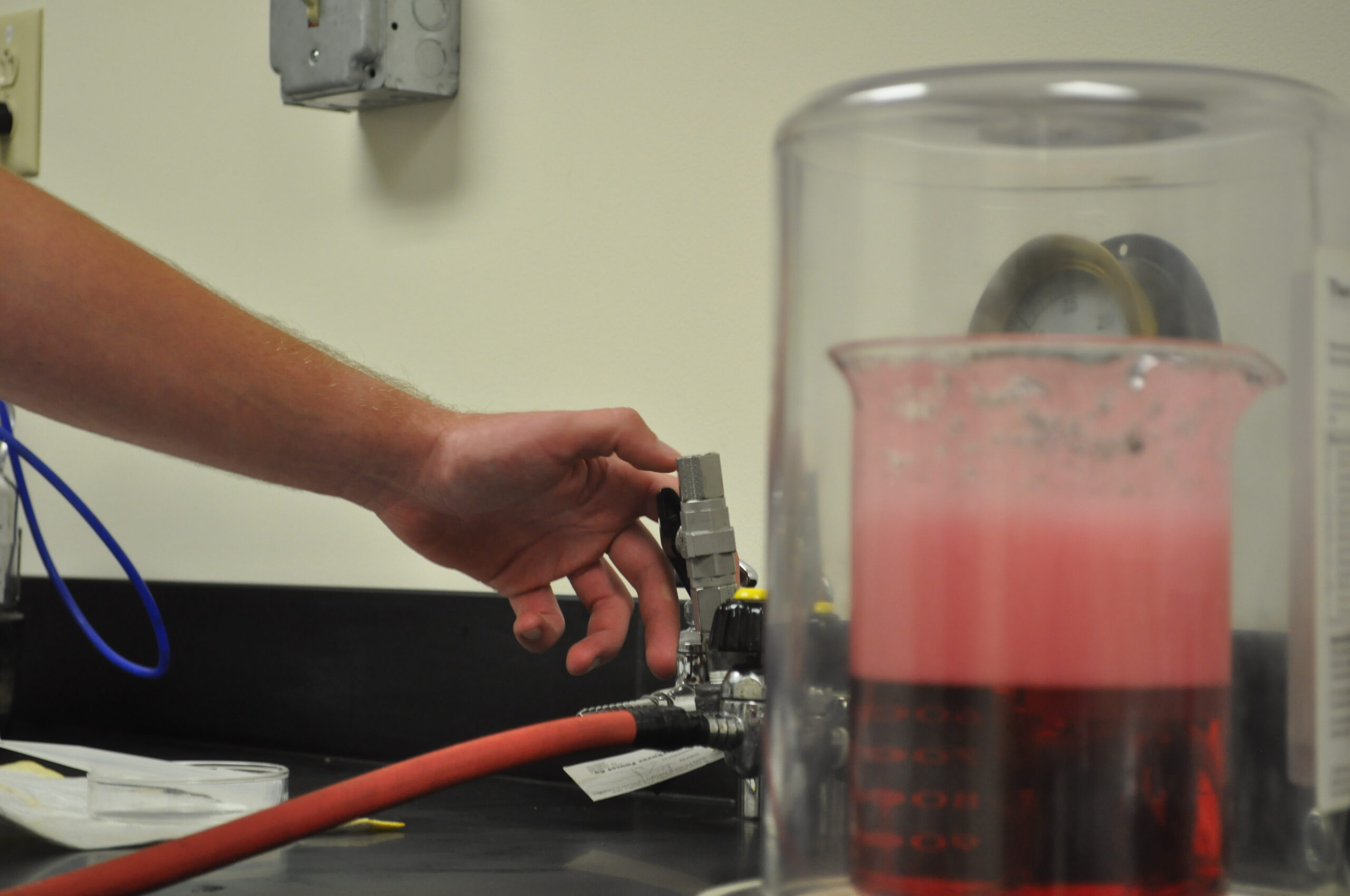 Hand adjusting a valve connected to a fluid analysis system, with a beaker containing a pink liquid in the foreground, illustrating the process of fluid sample analysis in a laboratory setting.
