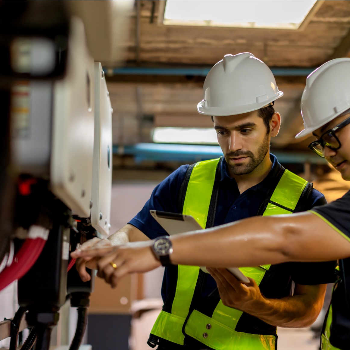 Two engineers in safety helmets and vests collaborate on machinery maintenance, examining equipment and reviewing data on a tablet in an industrial setting.