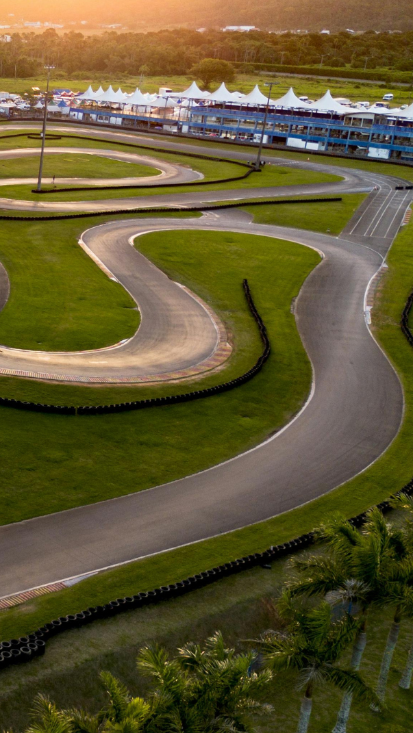 Aerial view of a karting track featuring winding curves and green grass, with a modern facility in the background and mountains on the horizon during sunset.