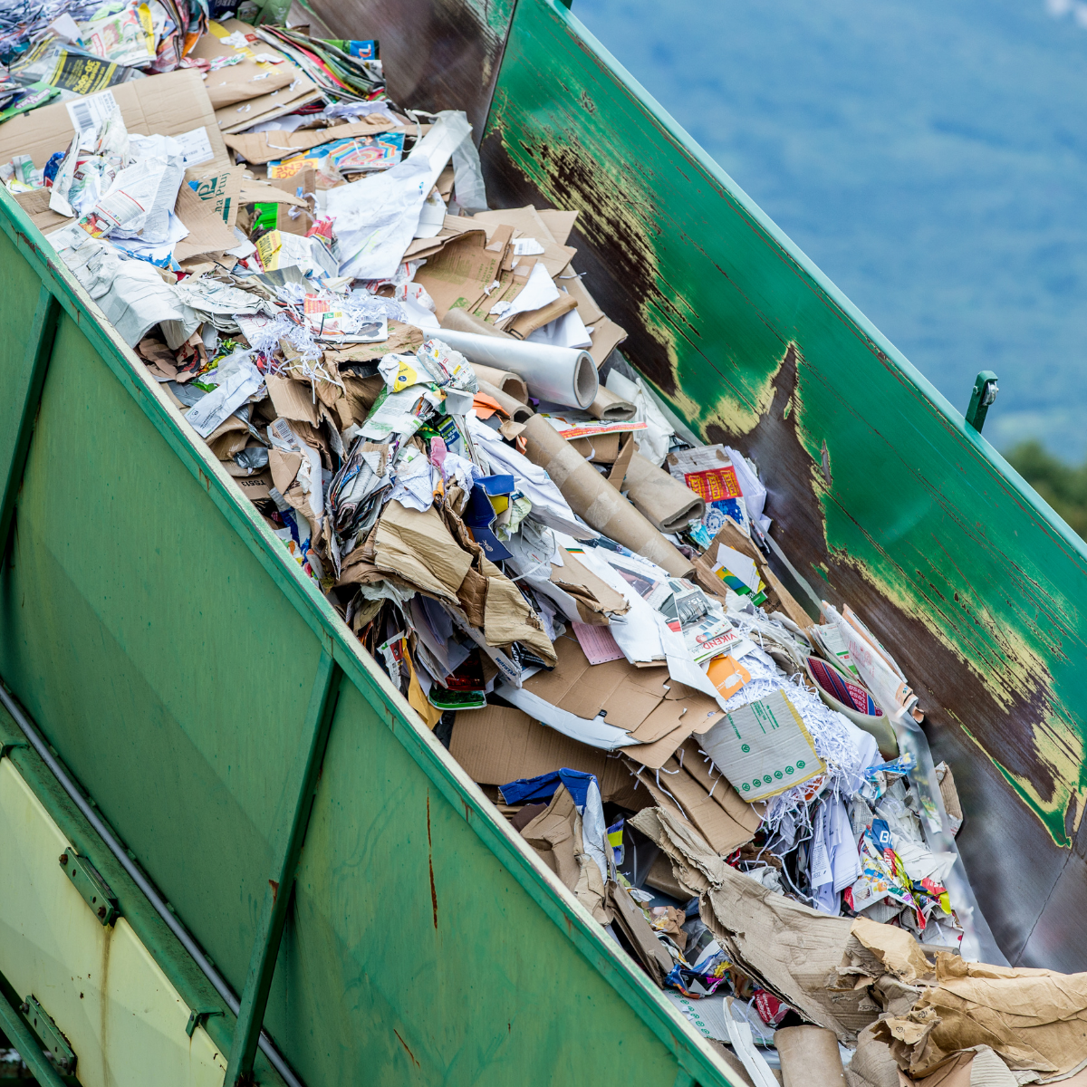 A close-up view of a green dumpster filled with various types of discarded paper and cardboard waste, showcasing the accumulation of recyclables at a landfill site.