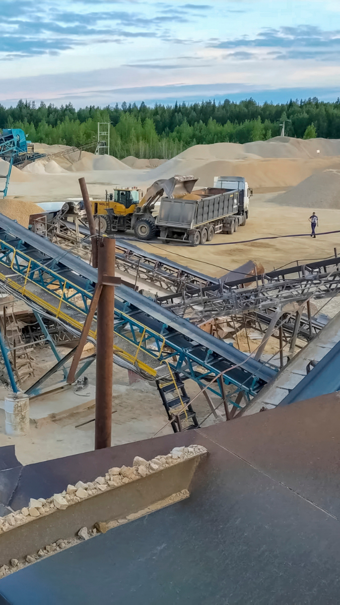 Aerial view of a material handling site featuring conveyor belts transporting aggregates, heavy machinery including a yellow loader and trucks, and a backdrop of natural landscape with trees and sand piles.