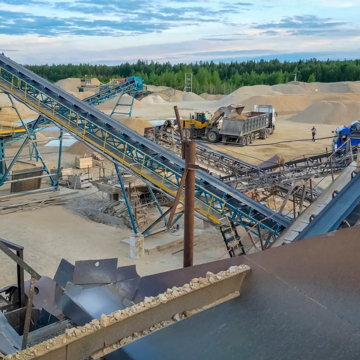 Aerial view of a material handling site featuring conveyor belts transporting aggregates, heavy machinery including a yellow loader and trucks, and a backdrop of natural landscape with trees and sand piles.