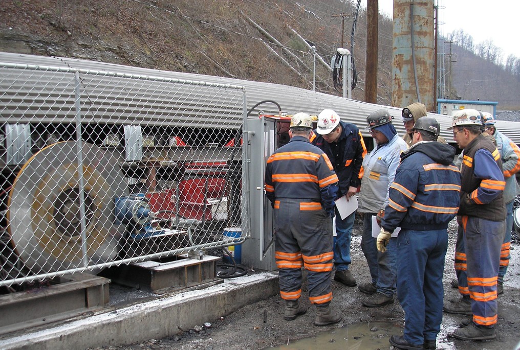 A group of field service technicians in safety gear discussing equipment operation near a fenced machinery setup, highlighting teamwork in industrial maintenance and repair.