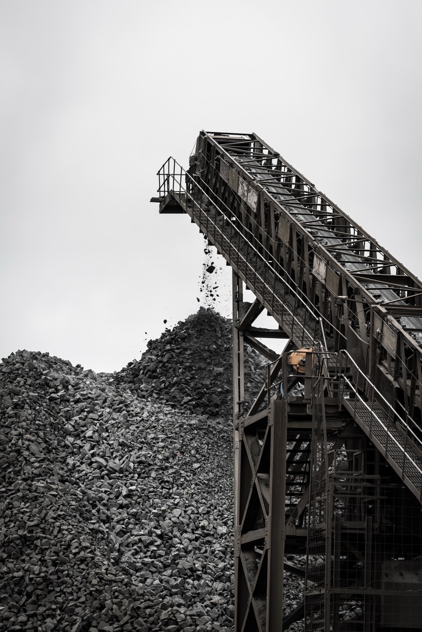 Conveyor belt transporting mined material, with rocks spilling onto a large pile below, showcasing industrial mining operations.