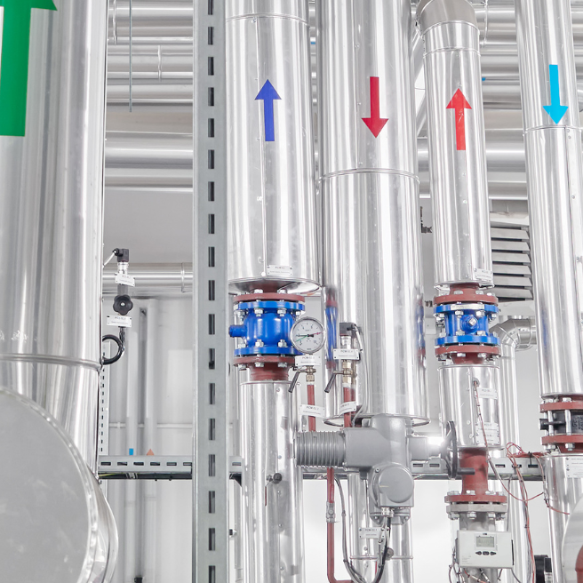 Industrial piping system in a renewable energy facility, featuring stainless steel pipes with colored directional arrows indicating flow direction, along with pressure gauges and valves for efficient energy management.