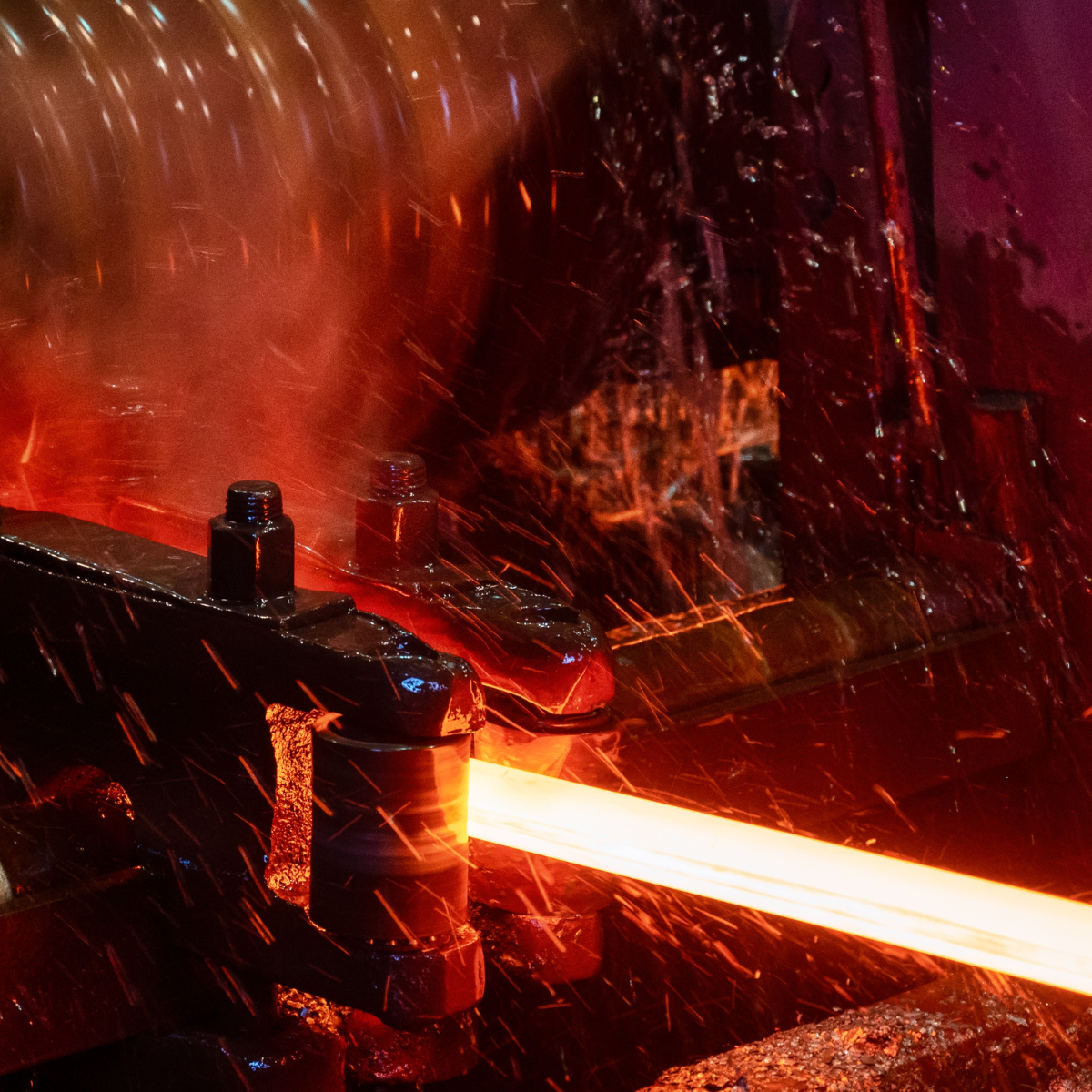 Close-up of a rolling mill process, showcasing a glowing red steel bar being shaped under high pressure, with water spray cooling the heated metal. The image highlights the intricate machinery and dynamic motion involved in metalworking.