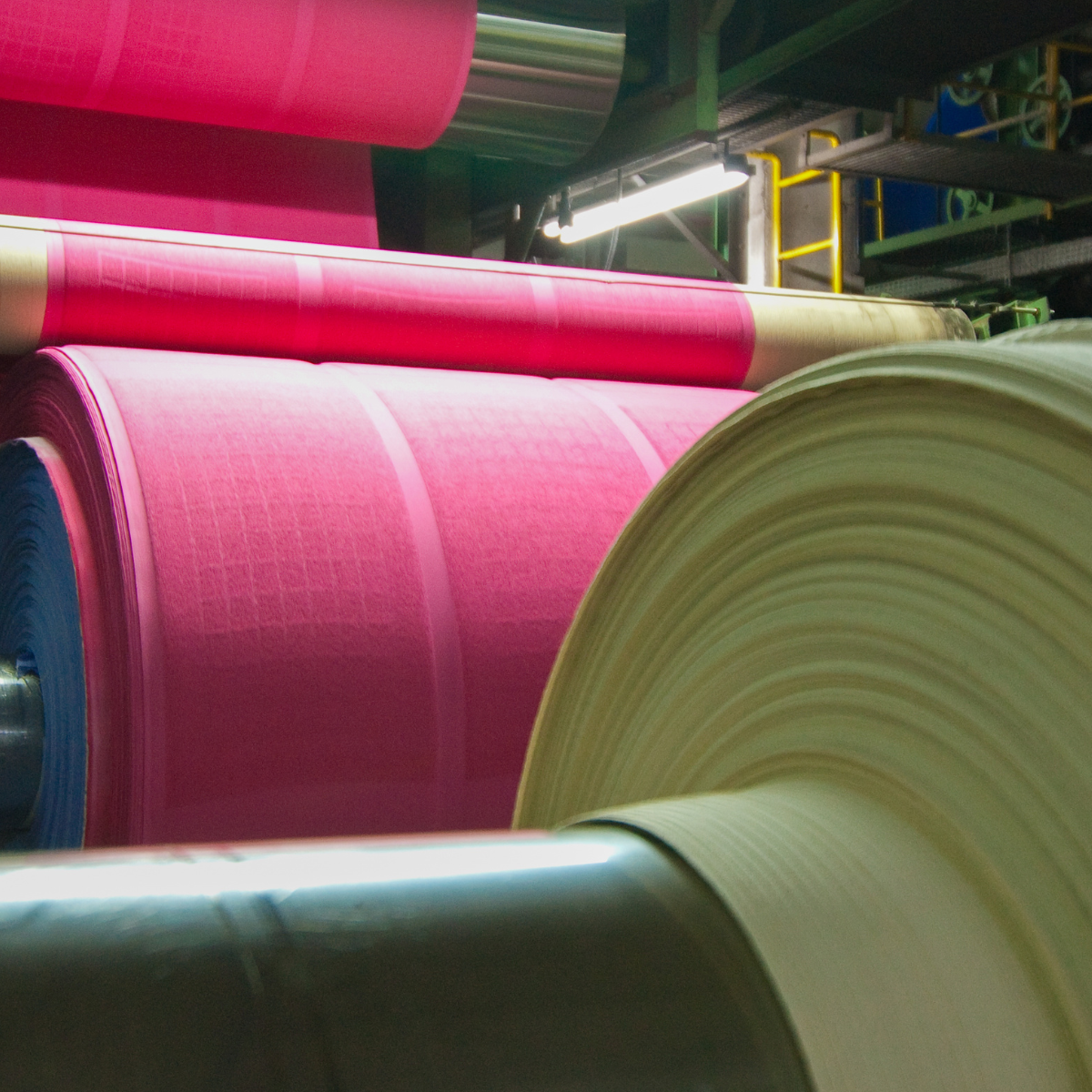 Close-up of large rolls of colorful paper in a manufacturing facility, showcasing the tensioning process used in paper production. The image features vibrant pink and blue rolls, highlighting the intricate machinery involved in the tensioning stage.