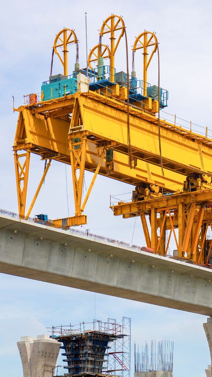 Yellow gantry crane positioned above a concrete bridge span, showcasing construction of a train rail system with supporting pillars and scaffolding in the background.