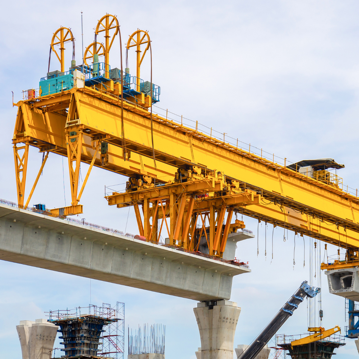 Yellow gantry crane positioned above a concrete bridge span, showcasing construction of a train rail system with supporting pillars and scaffolding in the background.
