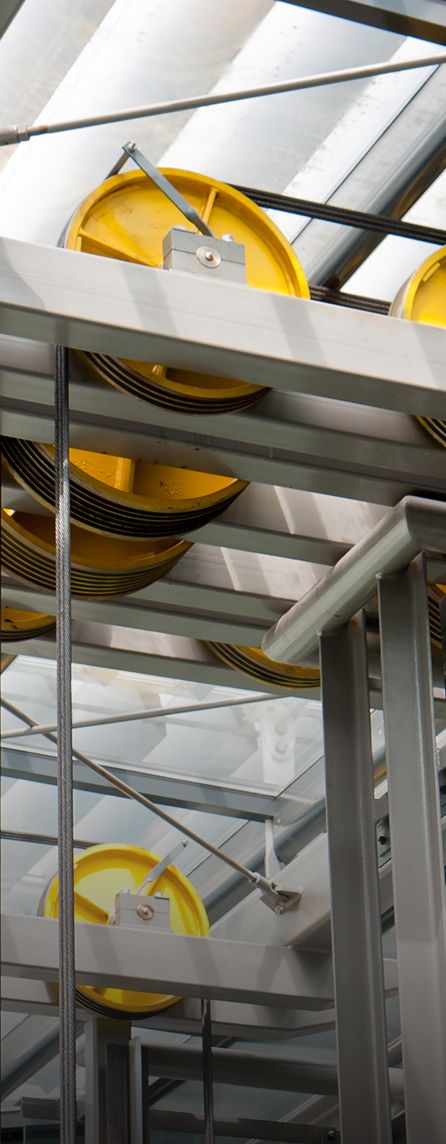 Industrial pulley system with yellow wheels and cables, mounted on a metal frame under a glass roof, showcasing mechanical engineering in a modern facility.